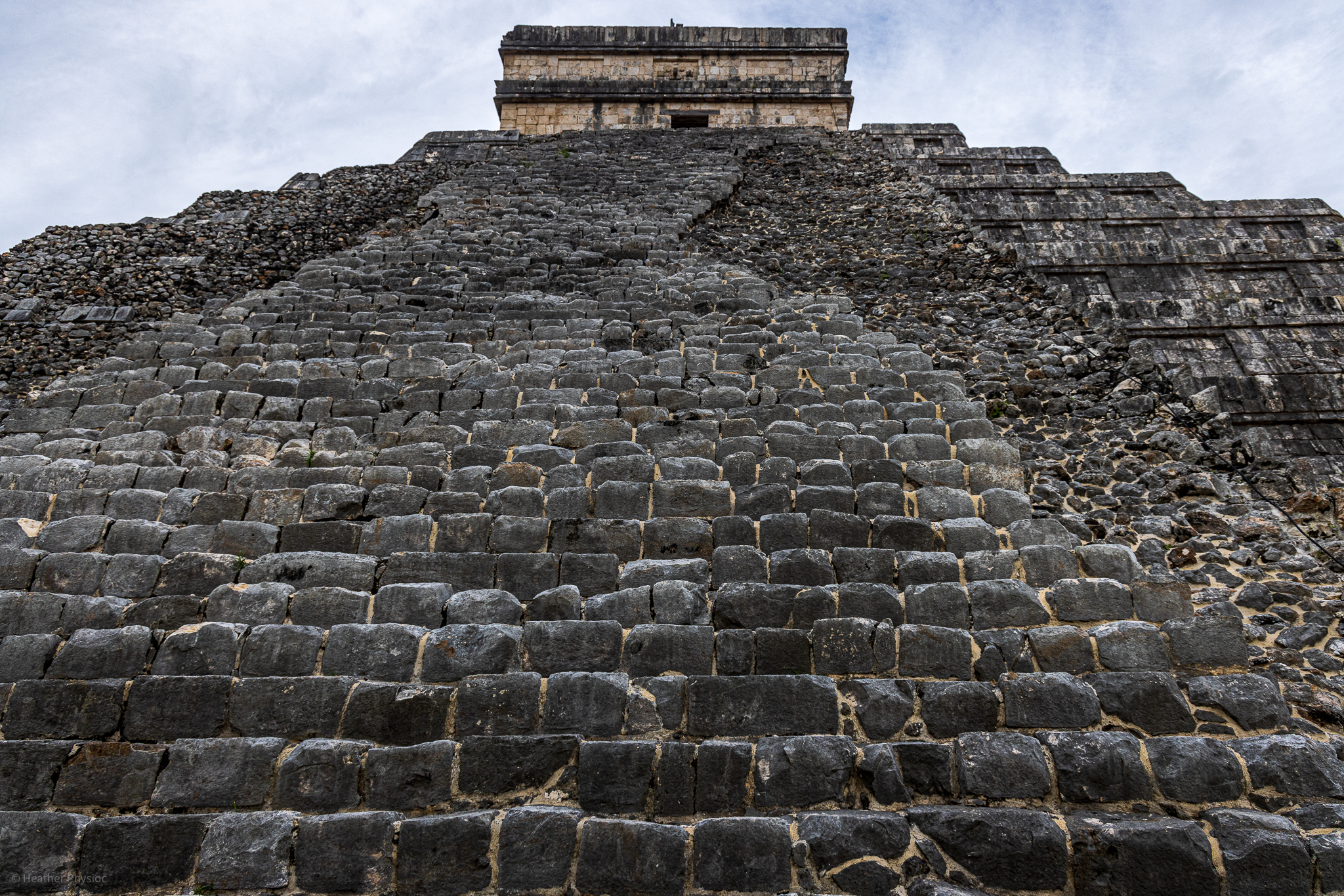 Upward view of the steep stairway of the Kukulkan Pyramid at Chichen Itza, showcasing the massive stone blocks and rugged texture, with the temple's peak looming against a cloudy sky.