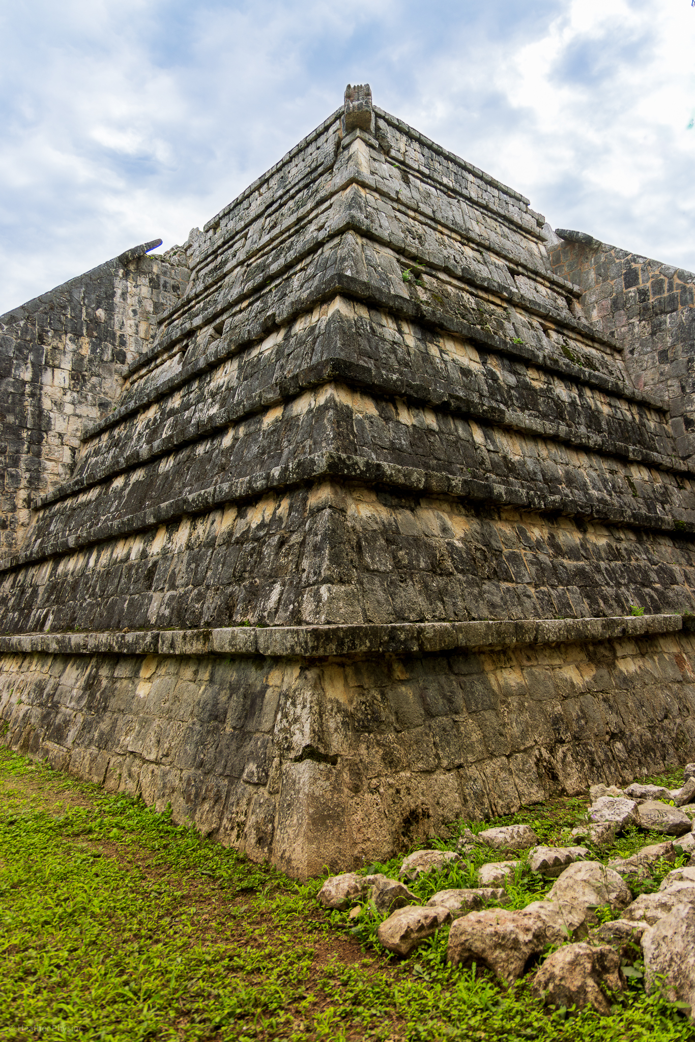The Ossuary, or High Priest's Grave, at Chichen Itza, seen from a low angle, highlighting the layered platforms and the temple's rounded corners against a soft cloudy sky, with green grass at its base.