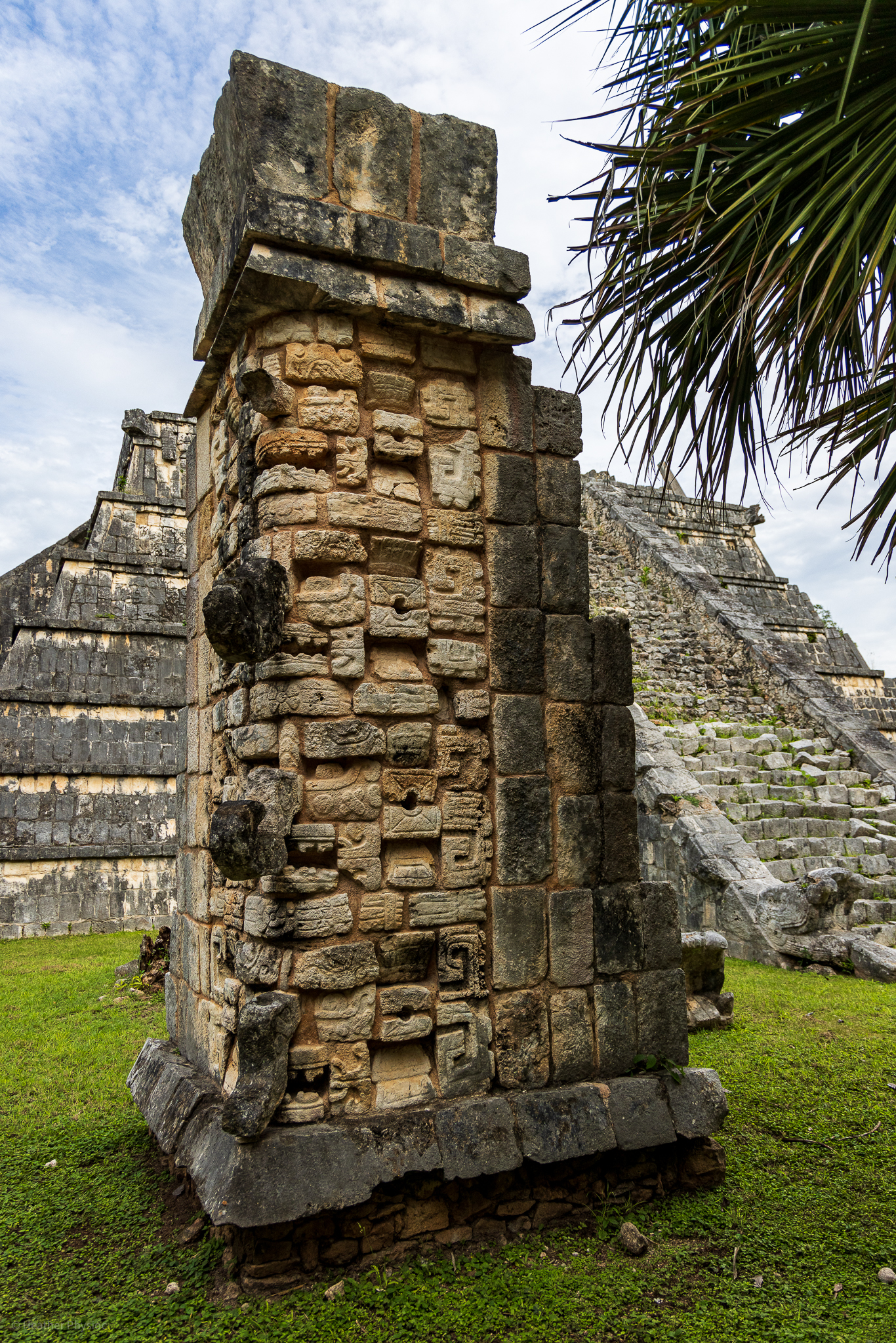 A detailed stone pillar with elaborate carvings, standing in front of the Kukulkan Pyramid at Chichen Itza. The foreground focuses on the intricate Mayan designs and glyphs etched into the rock.